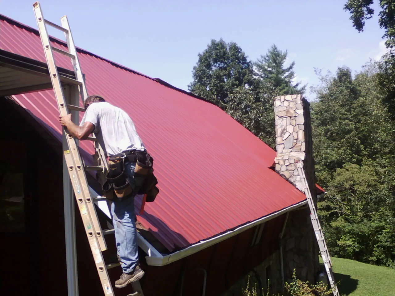 Roofer on a ladder beside a red metal roof and stone chimney — Soffit & Fascia Repair by Roofing Brothers in Mabel